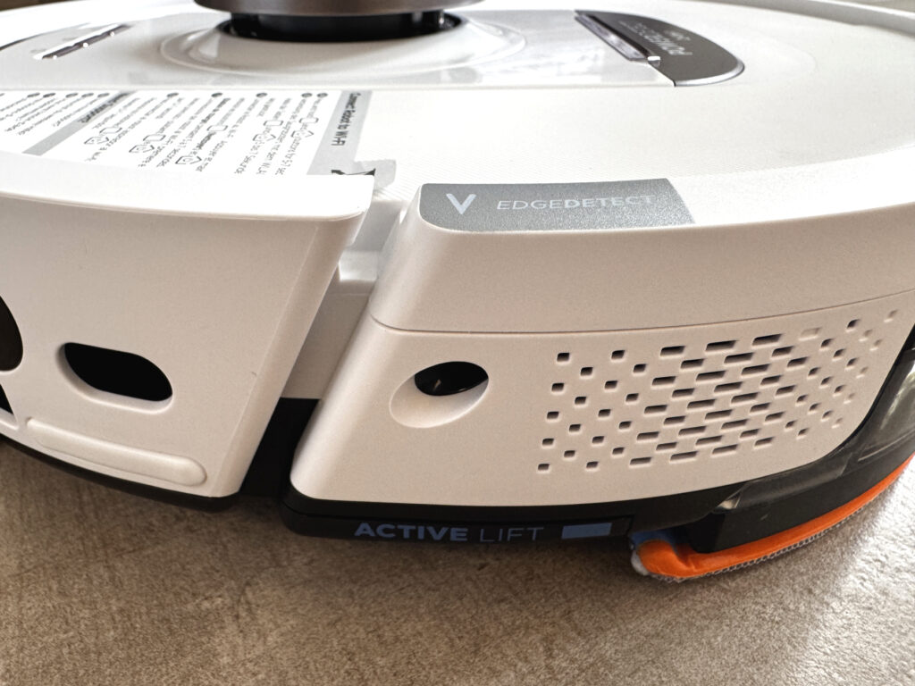 Nahaufnahme der Seitenansicht des Shark PowerDetect NeverTouch Pro 2-in-1. Sichtbar sind die EdgeDetect- und Active Lift-Funktionen. Die EdgeDetect-Technologie soll Schmutz aus Ecken effizienter entfernen, während Active Lift das Hinterteil des Roboters anhebt, um Teppiche beim Wischen trocken zu halten. Das Wischpad ist teilweise sichtbar, mit orangefarbenem Rand.